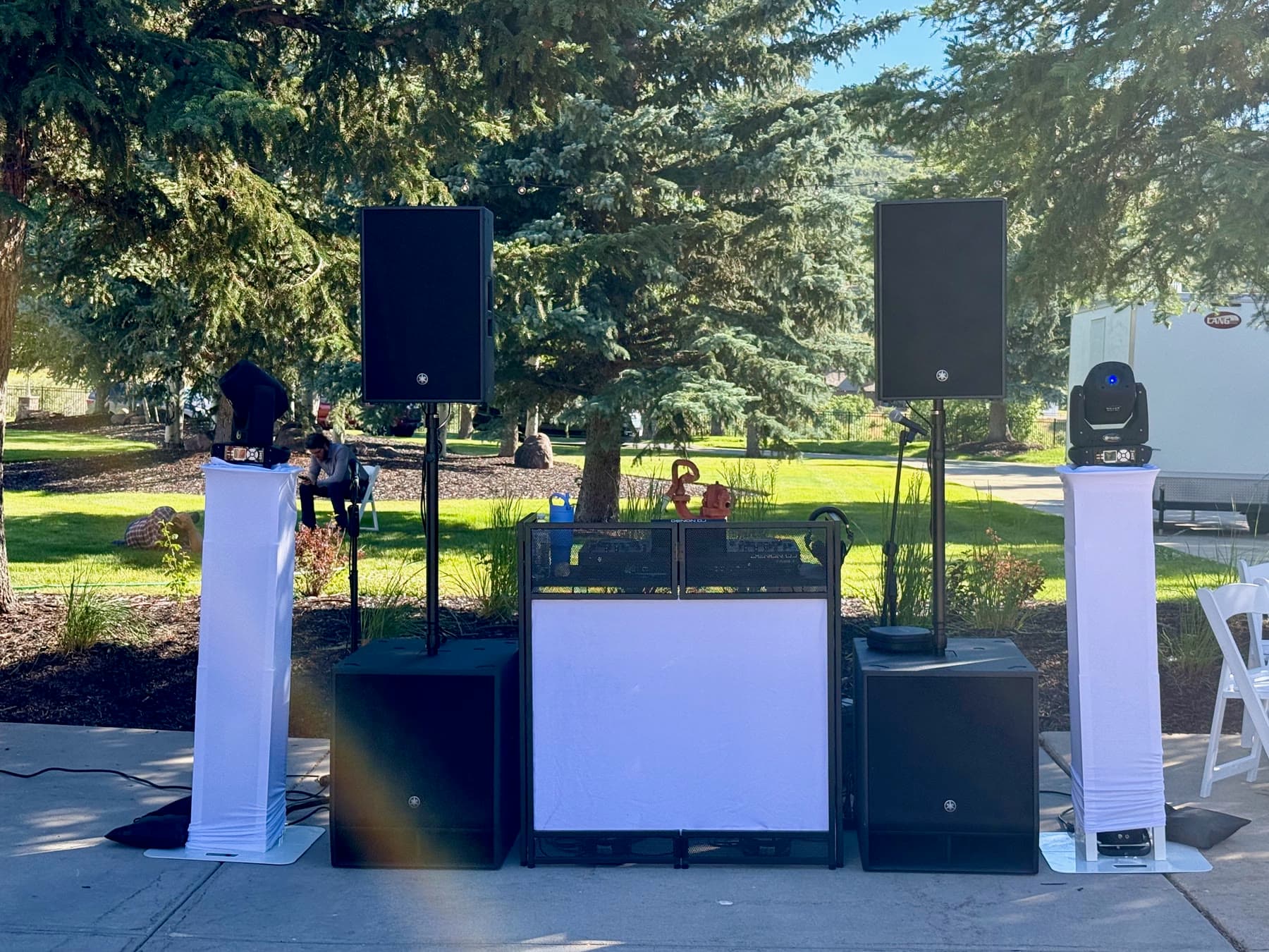 Outdoor wedding DJ setup with speakers, subs, booth lighting, and mover columns at a mountain venue