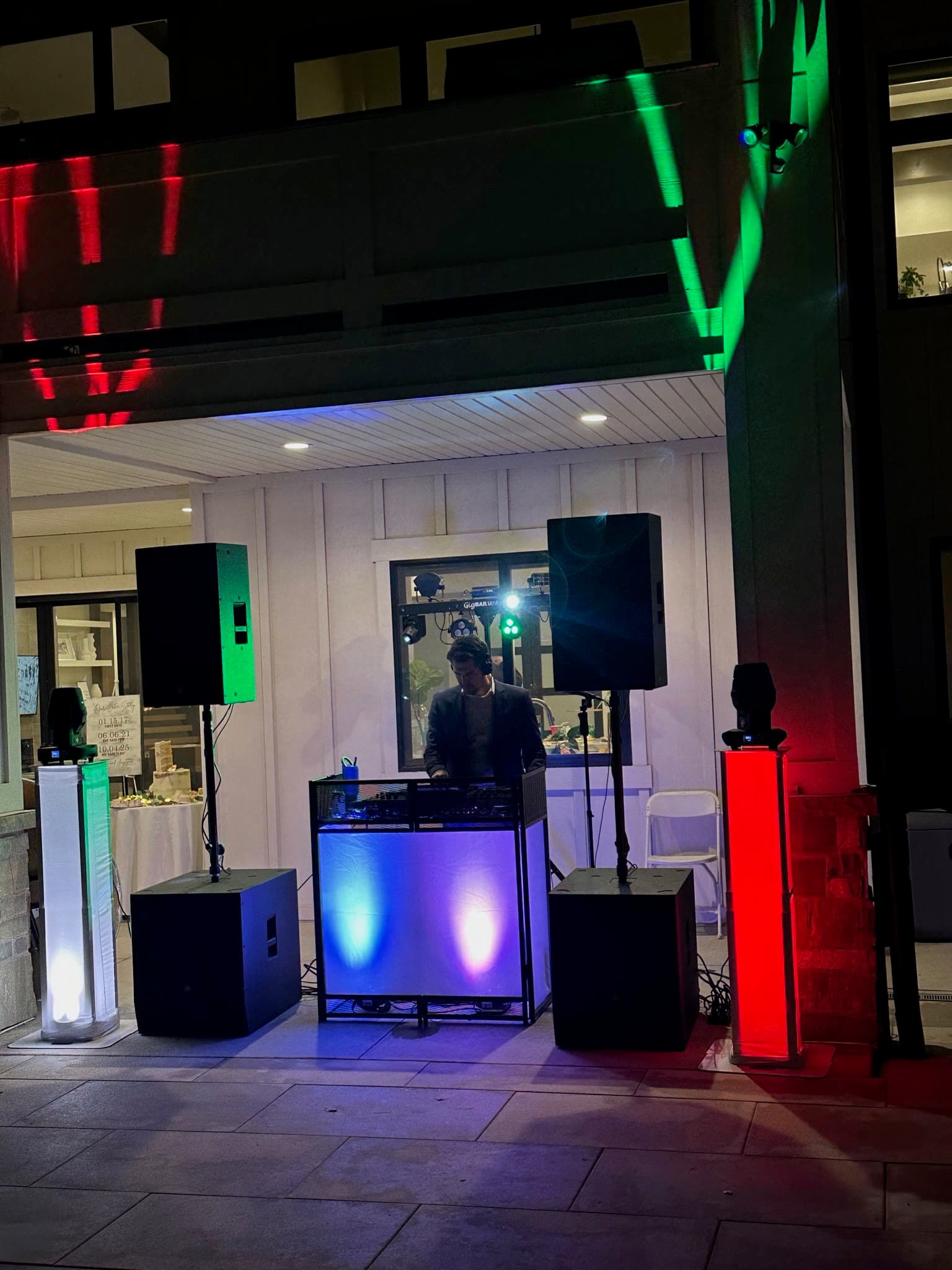 Outdoor reception DJ setup at night with speakers, booth lighting, and color-lit columns